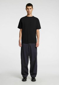 Juoda medvilninė trumpomis rankovėmis palaidinė, derinama su laisvai krentančiomis tamsiai mėlynomis kelnėmis ir juoda avalyne, stovinti priešais šviesią foną.