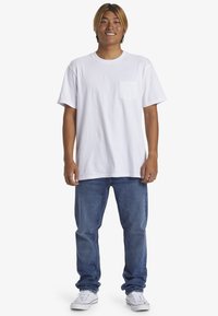 Biele bavlnené tričko s krátkym rukávom a vreckom, v kombinácii s modrými džínami. Model má na sebe biele tenisky a stojí pred jednoduchým pozadím.