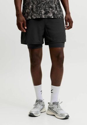 Homme portant un short noir superposé, des chaussettes blanches montantes, des chaussures de course grises et blanches, et une partie d'une chemise à motif noir et blanc.