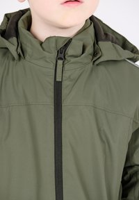 Giacca verde oliva con colletto alto, zip frontale e leggero lucido. Accenti neri sulla zip e sul cappuccio per contrasto.