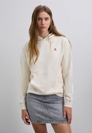 Sudadera con capucha color crema con un bolsillo delantero y un logo de calavera rojo, combinada con una falda vaquera gris con adornos de pedrería.