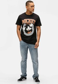 Camiseta negra con un gráfico de Mickey Mouse, combinada con jeans azul claro y zapatillas negras con franjas blancas. Diseño casual.