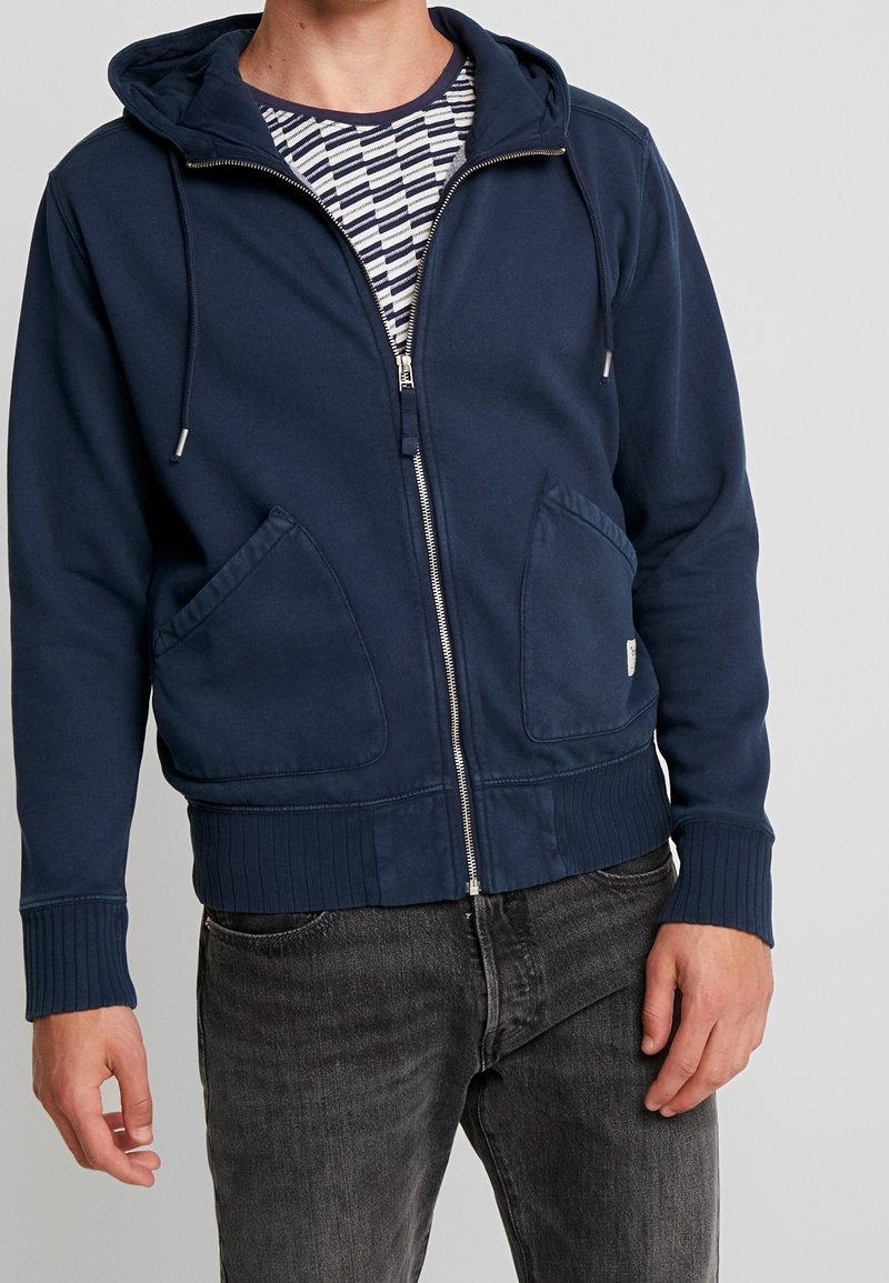 Sudadera con cremallera en color navy, con bolsillos tipo canguro, puños y dobladillo acanalados. Se lleva sobre una camiseta de rayas y cuenta con una capucha ajustable.