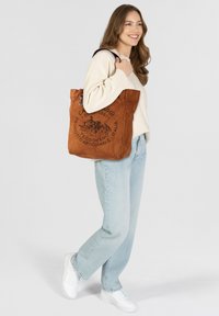 Bruine suède shopper met een bedrukte ontwerp met Italiaanse tekst, zilveren beslag en dubbele schouderbanden, gecombineerd met lichtblauwe spijkerbroek.