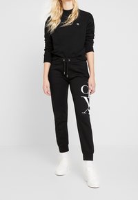 Sudadera de algodón negro con logo en el pecho, combinada con joggers negros que tienen un gráfico blanco. El modelo lleva zapatillas blancas.