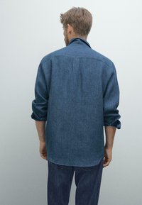 Uomo con le maniche arrotolate che indossa una camicia blu larga e jeans scuri, in piedi di fronte a uno sfondo grigio chiaro semplice.