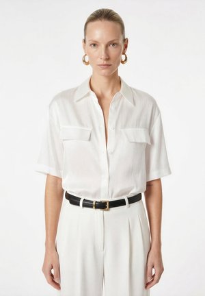 Femme aux cheveux blonds lissés en arrière, portant une chemise blanche à manches courtes boutonnée, un pantalon blanc, une ceinture noire et de grandes boucles d'oreilles dorées en forme d'anneau, regardant vers l'avant.
