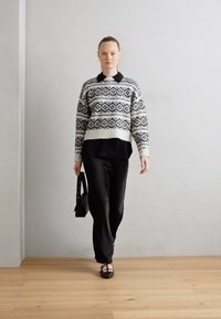 Jersey de punto en color crema con patrones geométricos negros, superpuesto sobre una camisa negra. Combinado con pantalones negros y bailarinas, sosteniendo un bolso de hombro.