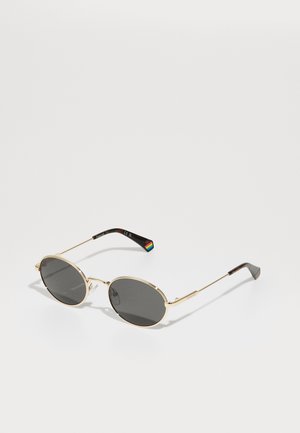 Goudkleurige ovale zonnebril met donkere lenzen. Dunne metalen steunen versierd met een regenboogdetail op de linkerkant. Gladde, glanzende afwerking.