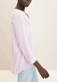 Maglietta a maniche lunghe rosa e bianca a righe con scollo tondo e tasca, realizzata in tessuto leggero. Jeans visibili sotto.