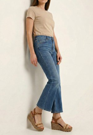 Kvinde læner sig op ad væggen iført en beige kortærmet top, korte blå jeans og brun kilehælssandaler med ankelsnore.