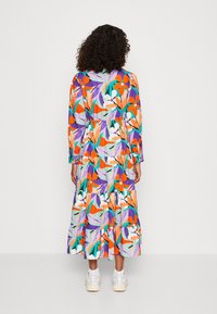 Robe colorée à manches longues avec un design à niveaux, présentant des motifs floraux orange, violet et vert. Portée avec des baskets blanches.