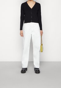 Persona vestida con un cárdigan negro de canalé con botones, pantalones rectos blancos, zapatos negros gruesos, sosteniendo un pequeño bolso verde con cadena dorada.