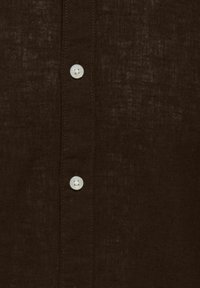 Tissu brun foncé texturé avec deux boutons blancs cousus alignés verticalement sur une patte de chemise.