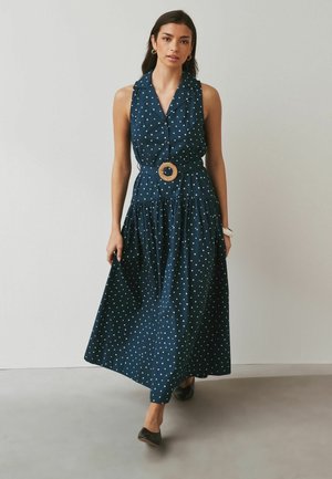 Kobieta w granatowej sukience w grochy bez rękawów z okrągłą plecioną klamrą paska, idąca do przodu na tle jednolitej szarej ściany.