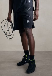 Idrettsutøver iført svarte shorts og sko, som holder et sammenvridd hoppetau, stående på en enkel gulv mot en nøytral vegg.