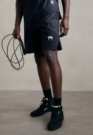 Athlete wearing black sports shorts and shoes holding a coiled jump rope, standing on a plain floor against a neutral wall.