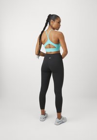 Turkise sports-BH med utskåret bakdesign kombinert med svarte leggings. Modell med grå treningssko. Glatt stofftekstur.