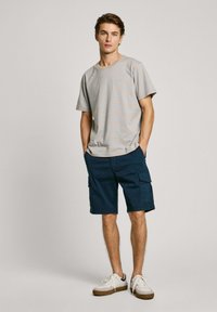 Pilkas ir mėlynas dryžuotas trumpas rankovės marškinėliai derinami su tamsiai mėlynomis krovinių šortais. Modelis dėvi baltus sportbačius, pabrėždamas kasdienišką aprangą.