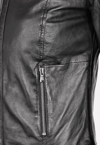 Gros plan d'une veste en cuir noire montrant une poche zippée verticale argentée sur le côté avec des coutures détaillées et une surface en cuir texturée.