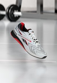 Λευκό, μαύρο και κόκκινο αθλητικό παπούτσι Reebok να αιωρείται πάνω από το δάπεδο γυμναστηρίου με θολές αλτήρες στο φόντο.