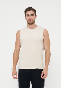 Άνδρας με μπεζ αμάνικη μπλούζα και μαύρο παντελόνι στέκεται μπροστά σε απλό λευκό φόντο, με το πρόσωπο προς τα εμπρός.