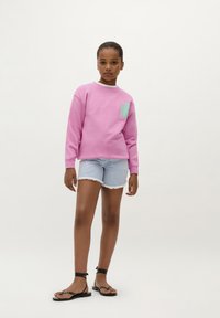 Felpa rosa con un piccolo design di tasca verde menta, abbinata a pantaloncini in denim azzurro chiaro e sandali neri piatti.