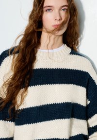 Krémový a námořnicky modrý pruhovaný pletený svetr s hrubou texturou, žebrovaným výstřihem a volným střihem, zobrazující vertikální a horizontální pruhy.