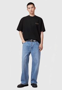 Μαύρο oversized T-shirt με μικρό χρυσό λογότυπο, συνδυασμένο με ανοιχτό μπλε τζιν παντελόνι με ίσια μάτια και μαύρα υφασμένα παπούτσια. Το μοντέλο στέκεται με τα χέρια στις τσέπες.