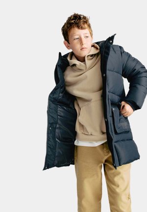 Granatowa kurtka puchowa o pikowanym wzorze, zapinana na zamek, z kieszeniami bocznymi, nałożona na beżowy sweatshirt z kapturem i jasne spodnie w kolorze khaki.