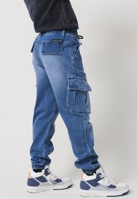 Kék farmer cargo jogger nadrág gumis zoknival és húzózsinóros derékkal. Nagy oldalsó zsebekkel és hátsó foltlogóval rendelkezik. Fehér és tengerészkék sportcipők.