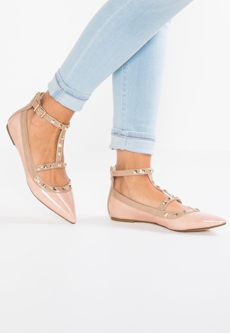 Pieds portant des ballerines pointues rose clair avec des brides cloutées, associées à un jean skinny bleu clair roulé sur un fond blanc.
