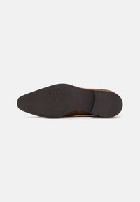 Talpa de pantof din suede maro, cu o talpă exterioară din cauciuc negru texturat. Prezintă o formă subțire și ascuțită, cu călcâi întărit pentru stabilitate.