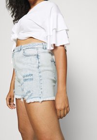 Vaaleansiniset korkeavyötäröiset farkkushortsit, joissa on kulutettuja yksityiskohtia, rispaantunut helma ja perinteinen viiden taskun malli. Yhdistetty valkoiseen röyhelöpaitaan.