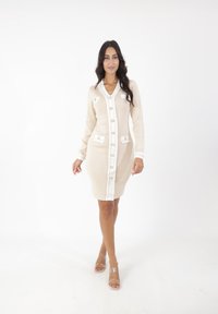 Abito in maglia beige con maniche lunghe, scollatura a V, chiusura anteriore con bottoni e due tasche. Il bordo bianco e i bottoni aggiungono contrasto. Indossato con tacchi trasparenti.