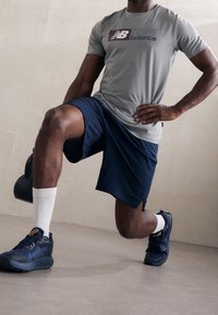 Szary t-shirt New Balance z logo, w połączeniu z granatowymi szortami. Czarne buty sportowe Gore-Tex, białe skarpetki do kostek i kettle w ręku.