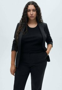 Blazer nero su misura sopra una maglia nera aderente a coste, abbinato a pantaloni aderenti coordinati. Texture liscia, design strutturato.