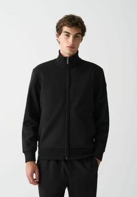 Fekete cipzáras dzseki magas gallérral, sima anyagból készült. Ráncolt mandzsettákkal és szegéllyel rendelkezik, minimalistának mondható dizájn, látható minták nélkül.