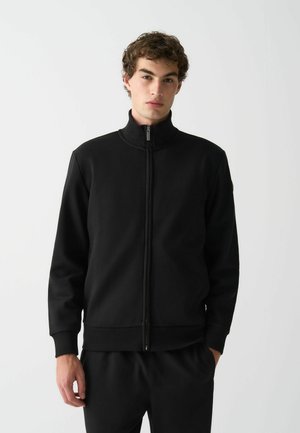 Veste noire zippée avec un col haut, en tissu lisse. Elle possède des poignets et un ourlet côtelés, avec un design minimaliste sans motifs visibles.
