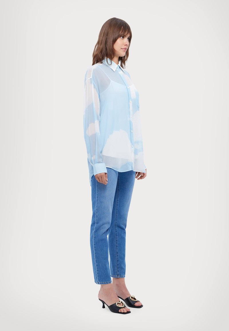 Chemise légère bleu clair envoûtante avec un motif de nuages, boutonnière à l'avant et col, associée à un jean bleu ajusté et des talons ouverts noirs avec des accents dorés.