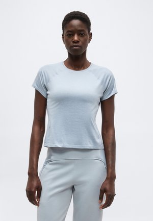 Žena s krátkymi vlasmi, ktorá má na sebe svetlomodré priliehavé tričko s krátkym rukávom a zodpovedajúce svetlomodré nohavice, stojaca pred jednofarebným pozadím.