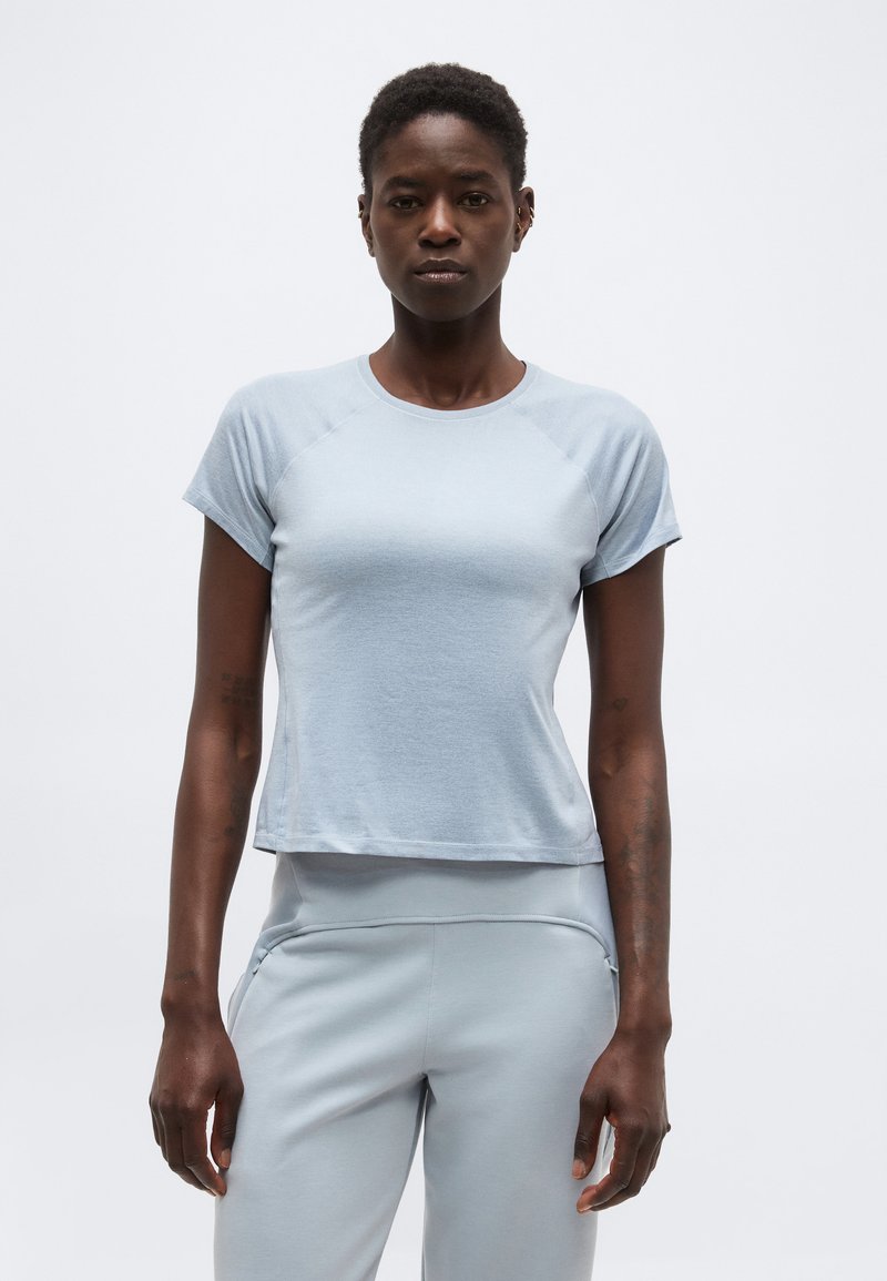 Γυναίκα με κοντά μαλλιά που φοράει ένα γαλάζιο εφαρμοστό κοντομάνικο μπλουζάκι και αντίστοιχο γαλάζιο παντελόνι, στέκεται μπροστά σε μια απλή φόντο.