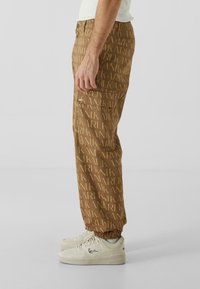 Pantaloni maro cu un model repetat al logo-ului "KARL", confecționați dintr-un material moale, cu o croială lejeră și manșete elastice, asortați cu adidași albi.