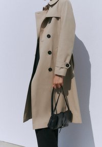 Trench-coat beige à double boutonnage, boutons noirs et col classique. Associé à un petit sac à main noir.