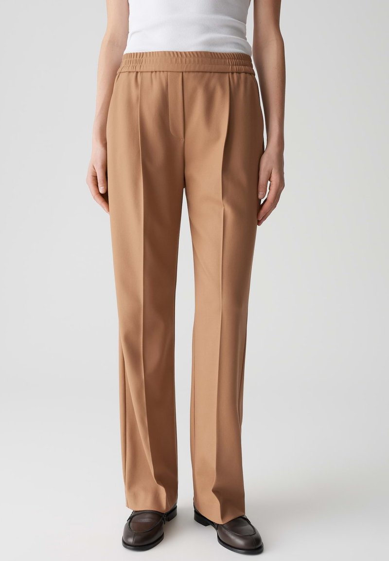 Pantaloni beige comodi con vita elastica e pieghe laterali. Tessuto liscio e design a gamba dritta, abbinati a scarpe scure.