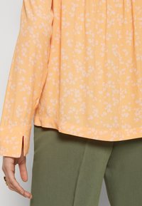 Blouse orange légère à motif floral avec un ourlet arrondi et des poignets fendus, associée à un pantalon vert. Texture douce, coupe décontractée.
