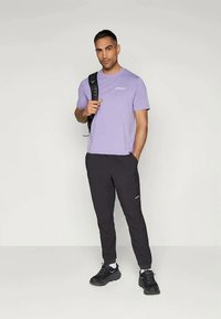 Lila Kurzarm-T-Shirt mit kleinem Logo, kombiniert mit schwarzen, schmal zulaufenden Jogginghosen und schwarzen Turnschuhen, dazu ein über eine Schulter geschulterter Rucksack.