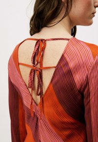 Blouse rayée rouge et rose avec un dos en V profond orné de liens décoratifs. Le tissu semble lisse avec des motifs fins verticaux et horizontaux.