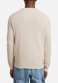 Maglione beige lavorato a maglia con texture a coste, scollo a giro e maniche lunghe. Presenta una piccola etichetta con logo vicino all'orlo, abbinato a jeans blu.
