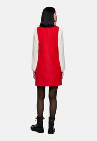Rotes ärmelloses Kleid mit strukturiertem Design, aus Stoff gefertigt. Über einem weißen Langarmoberteil getragen, kombiniert mit schwarzen Schnürstiefeln.